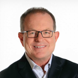 A middle-aged man with short hair, glasses, and a dark suit jacket smiles at the camera against a plain white background.